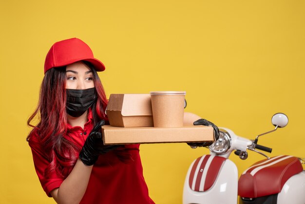 Vista frontale del corriere femminile in maschera con cibo di consegna sulla parete gialla