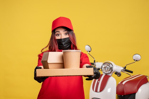 Vista frontale del corriere femminile in maschera con cibo di consegna sulla parete gialla