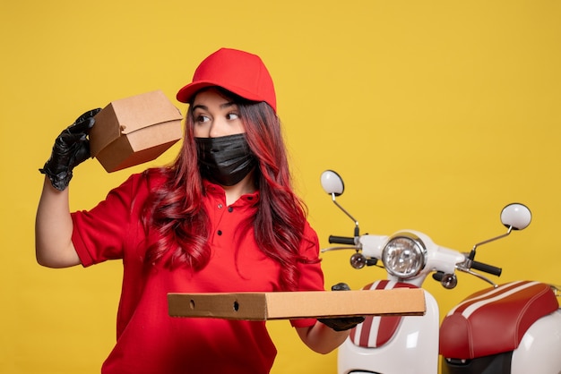 Vista frontale del corriere femminile in maschera con cibo di consegna sulla parete gialla