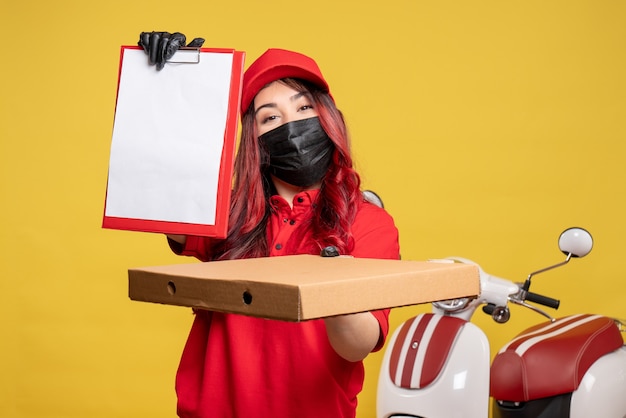 Vista frontale del corriere femminile in maschera con scatola di cibo di consegna e nota di file sulla parete gialla