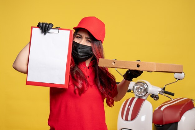 Vista frontale del corriere femminile in maschera con scatola di cibo di consegna e nota di file sulla parete gialla