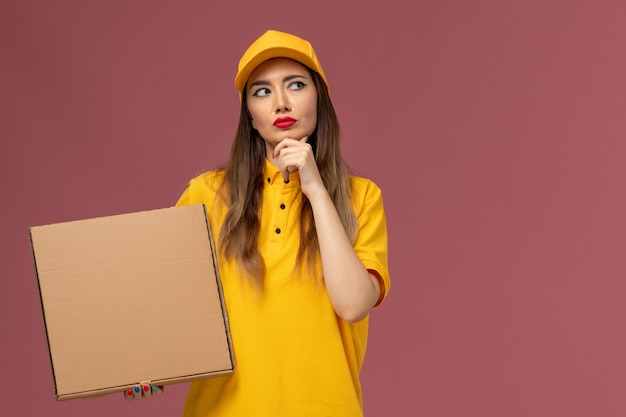 Vista frontale del corriere femminile in uniforme gialla e cappuccio che tiene il contenitore di cibo pensando sulla parete rosa chiaro