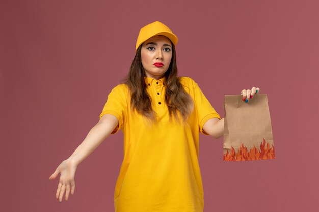 Vista frontale del corriere femminile in uniforme gialla e cappuccio che tiene il pacchetto alimentare con espressione ocnfused sulla parete rosa chiaro