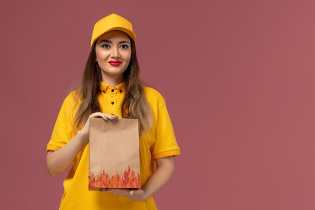 Vista frontale del corriere femminile in uniforme gialla e cappuccio che tiene il pacchetto alimentare e sorridente sulla parete rosa chiaro