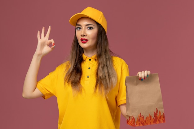 Vista frontale del corriere femminile in uniforme gialla e cappuccio che tiene il pacchetto alimentare sulla parete rosa chiaro