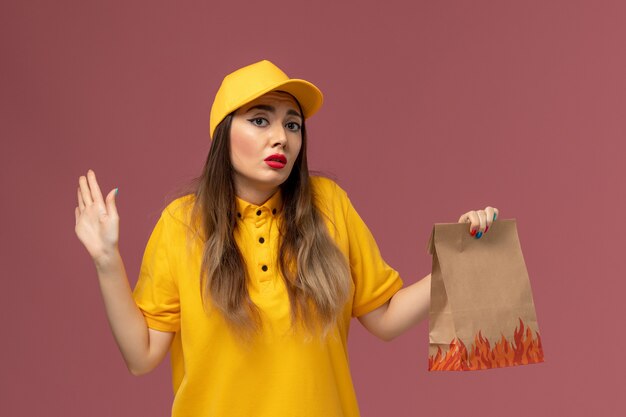 Vista frontale del corriere femminile in uniforme gialla e cappuccio che tiene il pacchetto alimentare sulla parete rosa chiaro