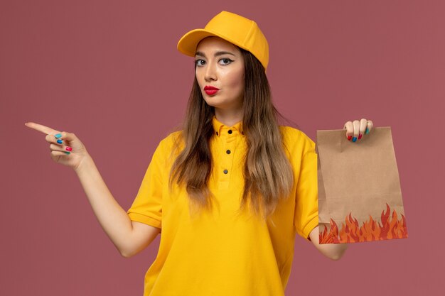 Vista frontale del corriere femminile in uniforme gialla e cappuccio che tiene il pacchetto alimentare sulla parete rosa chiaro