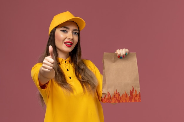 Vista frontale del corriere femminile in uniforme gialla e cappuccio che tiene il pacchetto alimentare sulla parete rosa chiaro