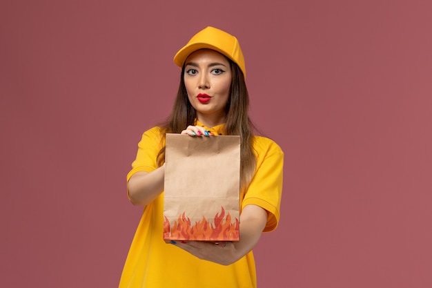 Vista frontale del corriere femminile in uniforme gialla e cappuccio che tiene il pacchetto alimentare sulla parete rosa chiaro