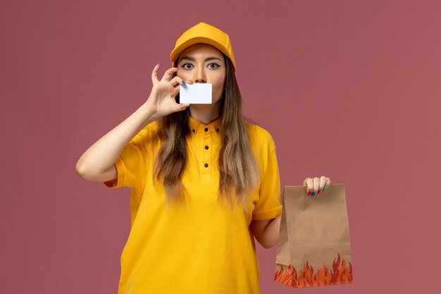 Vista frontale del corriere femminile in uniforme gialla e cappuccio che tiene il pacchetto di cibo e carta di plastica sulla parete rosa