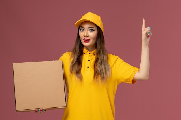 Vista frontale del corriere femminile in uniforme gialla e cappuccio che tiene la scatola di cibo sulla parete rosa chiaro