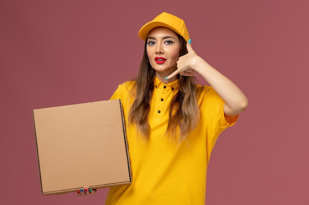 Vista frontale del corriere femminile in uniforme gialla e cappuccio che tiene la scatola di cibo sulla parete rosa chiaro