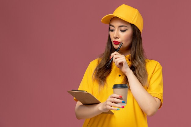 Vista frontale del corriere femminile in uniforme gialla e cappuccio che tiene la tazza di caffè di consegna e il blocco note con la penna sulla parete rosa