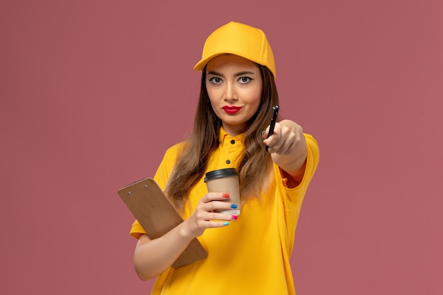 Vista frontale del corriere femminile in uniforme gialla e cappuccio che tiene la tazza di caffè di consegna e il blocco note con la penna sulla parete rosa