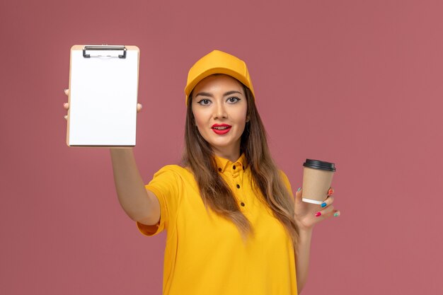 Vista frontale del corriere femminile in uniforme gialla e cappuccio che tiene la tazza di caffè di consegna e il blocco note sulla parete rosa