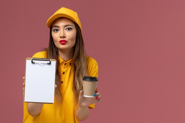 Vista frontale del corriere femminile in uniforme gialla e cappuccio che tiene la tazza di caffè di consegna e il blocco note sulla parete rosa