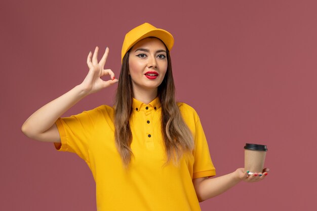 Vista frontale del corriere femminile in uniforme gialla e cappuccio che tiene la tazza di caffè di consegna sulla parete rosa