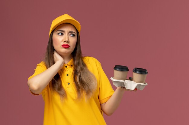 Vista frontale del corriere femminile in uniforme gialla e cappuccio che tiene le tazze di caffè di consegna che hanno mal di collo sulla parete rosa