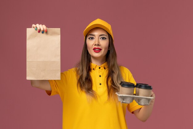 Vista frontale del corriere femminile in uniforme gialla e cappuccio che tiene le tazze di caffè di consegna marroni e il pacchetto di cibo sulla parete rosa