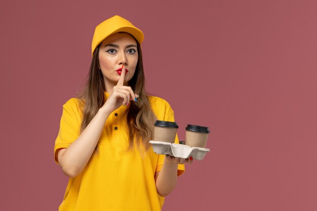 Vista frontale del corriere femminile in uniforme gialla e cappuccio che tiene le tazze di caffè di consegna sulla parete rosa