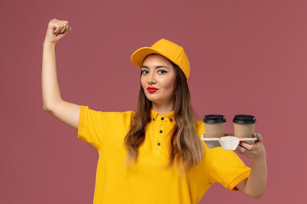 Vista frontale del corriere femminile in uniforme gialla e cappuccio che tiene le tazze di caffè di consegna sulla parete rosa