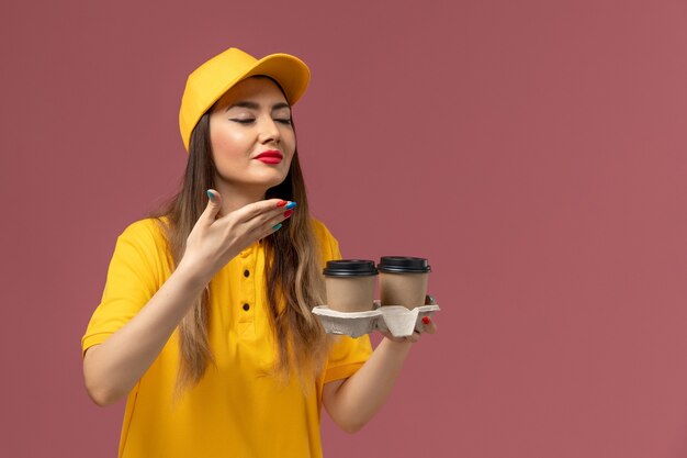 Vista frontale del corriere femminile in uniforme gialla e cappuccio che tiene le tazze di caffè di consegna sulla parete rosa