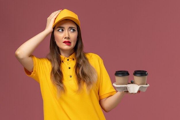 Vista frontale del corriere femminile in uniforme gialla e cappuccio che tiene le tazze di caffè di consegna sulla parete rosa