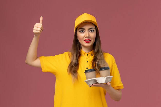 Vista frontale del corriere femminile in uniforme gialla e cappuccio che tiene le tazze di caffè di consegna sulla parete rosa