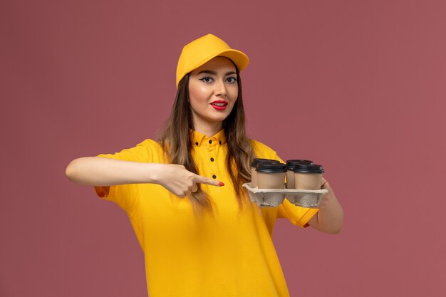 Vista frontale del corriere femminile in uniforme gialla e cappuccio che tiene le tazze di caffè marroni di consegna sulla parete rosa