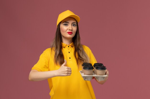 Vista frontale del corriere femminile in uniforme gialla e cappuccio che tiene le tazze di caffè marroni di consegna sulla parete rosa