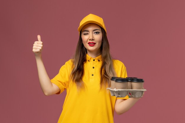 Vista frontale del corriere femminile in uniforme gialla e cappuccio che tiene le tazze di caffè marroni di consegna sulla parete rosa