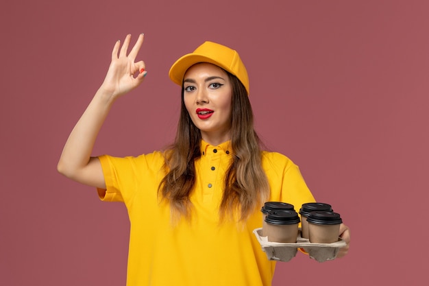 Vista frontale del corriere femminile in uniforme gialla e cappuccio che tiene le tazze di caffè marroni di consegna sulla parete rosa