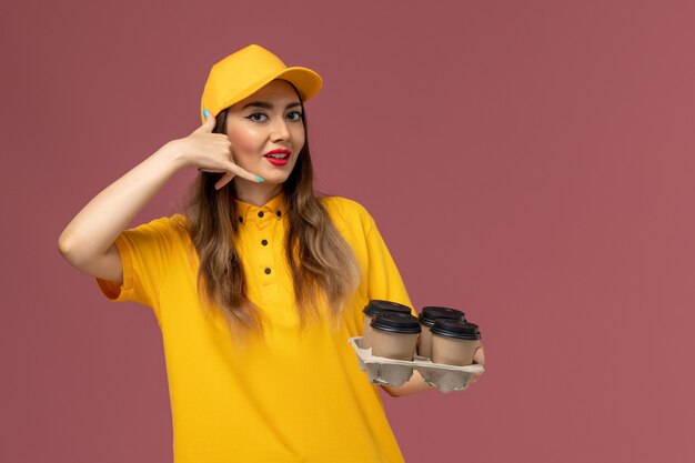 Vista frontale del corriere femminile in uniforme gialla e cappuccio che tiene le tazze di caffè marroni di consegna sulla parete rosa