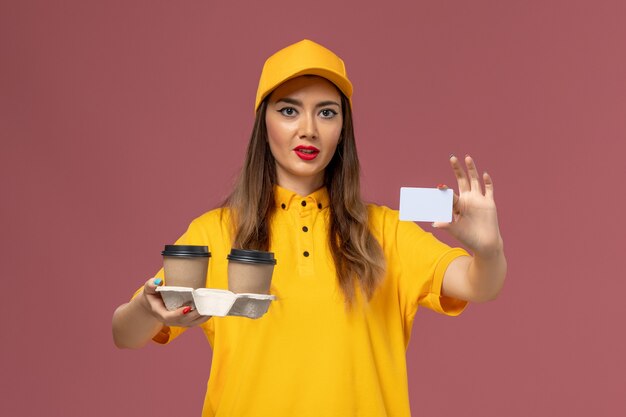 Vista frontale del corriere femminile in uniforme gialla e cappuccio che tiene le tazze e la carta di caffè di consegna sulla parete rosa