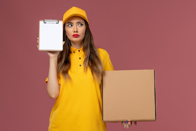 Vista frontale del corriere femminile in uniforme gialla e cappuccio che tiene scatola di cibo e blocco note pensando sulla parete rosa