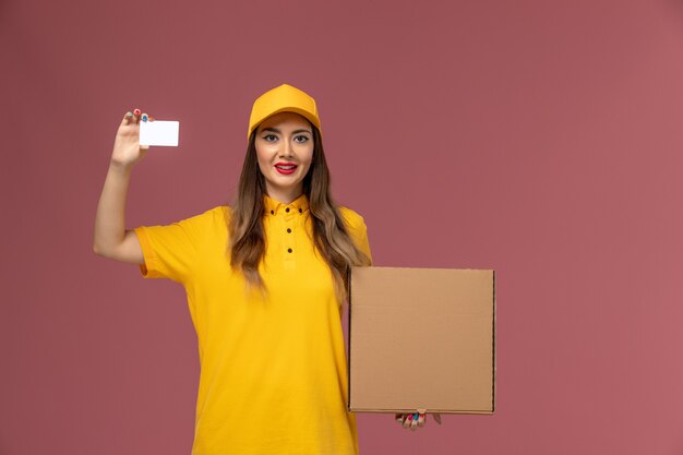 Vista frontale del corriere femminile in uniforme gialla e cappuccio che tiene scatola di cibo e carta di plastica sulla parete rosa chiaro