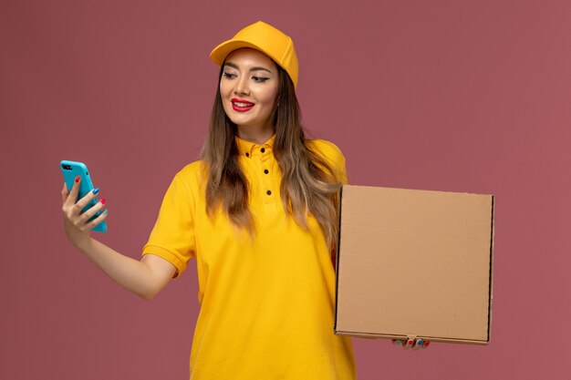 Vista frontale del corriere femminile in uniforme gialla e cappuccio che tiene scatola di cibo e telefono di lavoro sulla parete rosa chiaro