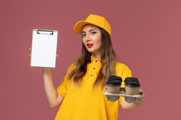 Vista frontale del corriere femminile in uniforme gialla e cappuccio che tiene tazze di caffè marroni e blocco note sulla parete rosa