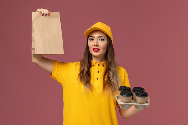 Vista frontale del corriere femminile in uniforme gialla e cappuccio che tiene tazze di caffè marroni e pacchetto di cibo sulla parete rosa