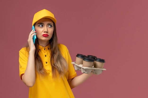 Vista frontale del corriere femminile in uniforme gialla e cappuccio che tiene tazze di caffè marroni e parla al telefono sulla parete rosa