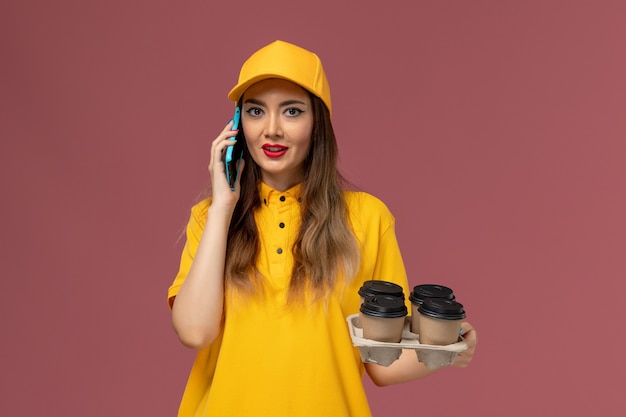 Vista frontale del corriere femminile in uniforme gialla e cappuccio che tiene tazze di caffè marroni e parla al telefono sulla parete rosa