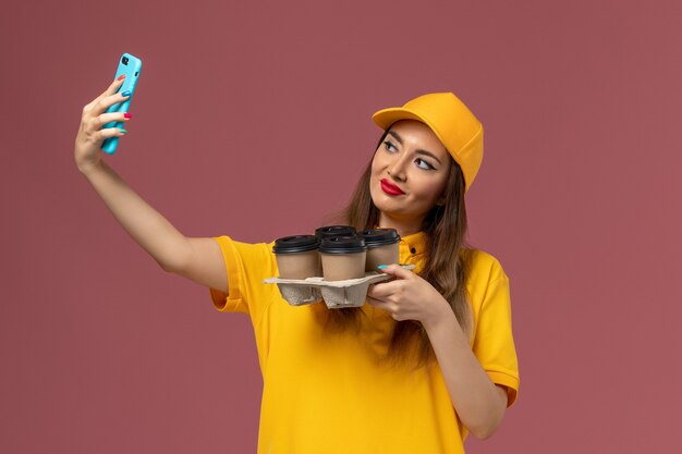 Vista frontale del corriere femminile in uniforme gialla e cappuccio che tiene tazze di caffè marroni e prendendo selfie sulla parete rosa