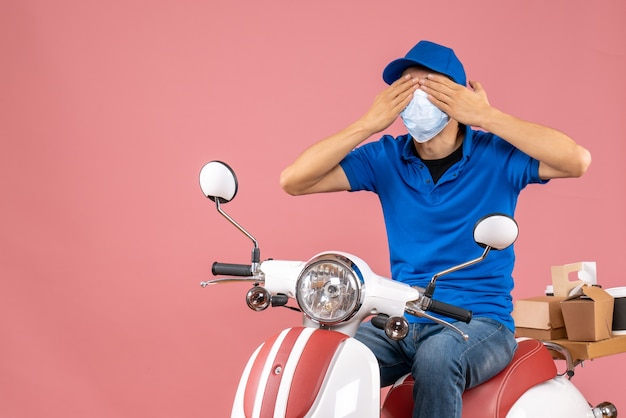 Vista frontale del corriere in maschera medica che indossa un cappello seduto su uno scooter chiudendo gli occhi su sfondo color pesca pastello