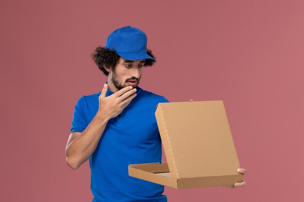 Vista frontale del corriere maschio in berretto blu uniforme con scatola di cibo aperta sulle mani sulla parete rosa chiaro