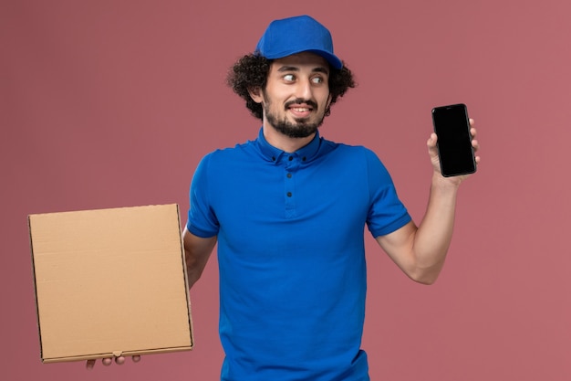 Vista frontale del corriere maschio in berretto blu uniforme con scatola di cibo e telefono sulle mani sul muro rosa chiaro