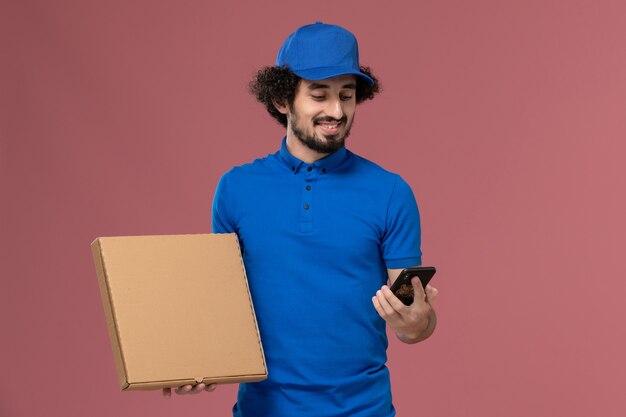 Vista frontale del corriere maschio in berretto blu uniforme con scatola di cibo e telefono sulle mani sul muro rosa chiaro