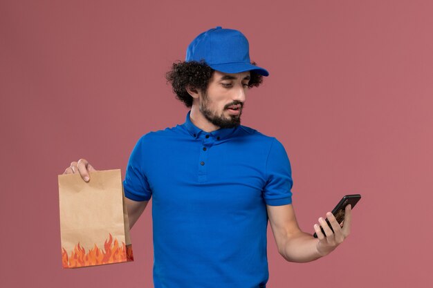 Vista frontale del corriere maschio in protezione uniforme blu con pacchetto di cibo di consegna sulle mani utilizzando il telefono sulla parete rosa