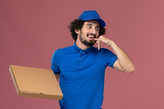 Vista frontale del corriere maschio in protezione uniforme blu con scatola di cibo sulle mani in posa sul muro rosa chiaro