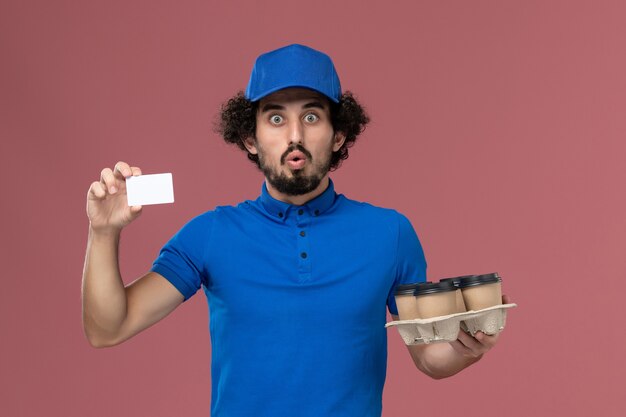 Vista frontale del corriere maschio in protezione uniforme blu con tazze di caffè di consegna e carta di plastica sulle mani sul muro rosa