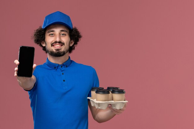 Vista frontale del corriere maschio in protezione uniforme blu con tazze di caffè di consegna sulle sue mani e il suo telefono di lavoro sulla parete rosa